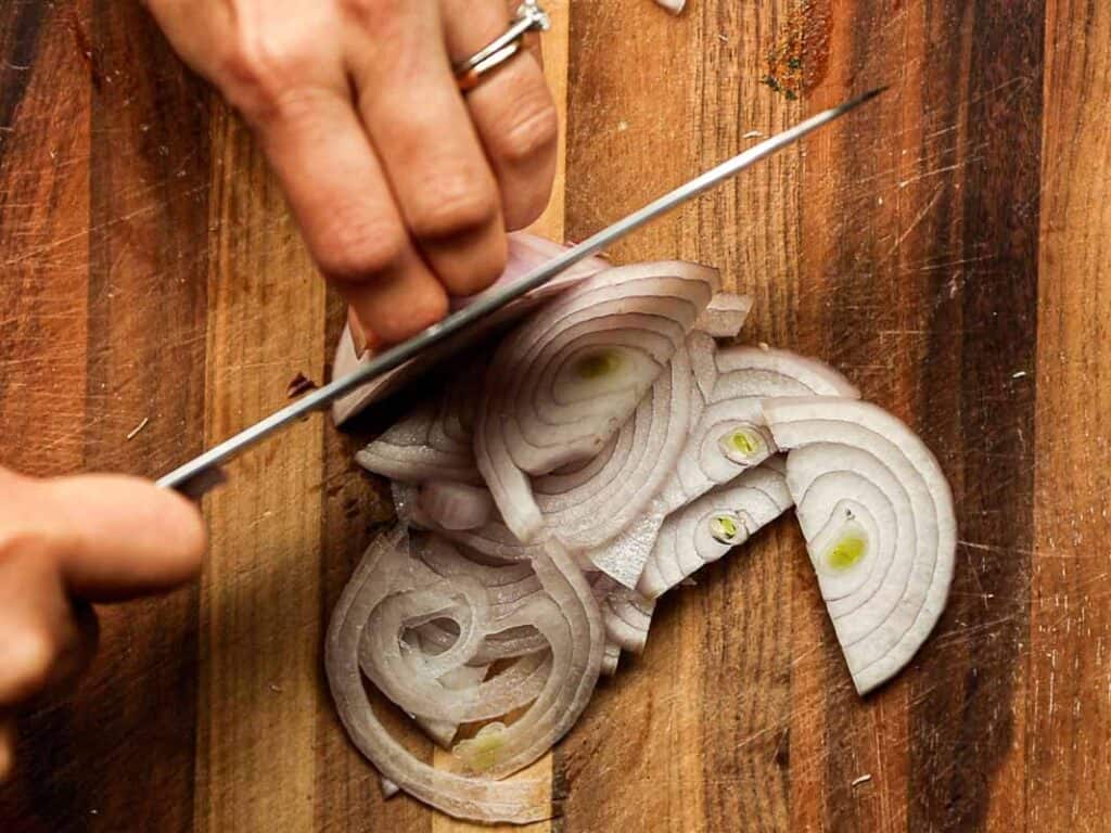 A close-up of a person slicing a shallot on a wooden cutting board. The shallot is cut into thin, circular slices. The person is wearing a ring and holding a knife with one hand, pressing down on the shallot with the other.